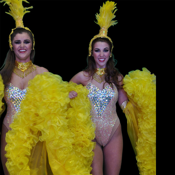 Two women modeling bright yellow carnival costumes with large ruffled fabric pieces, sequined bodysuits, feathered headpieces, and gold necklaces against a black background.